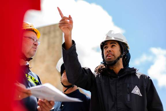A man in a hard hat is pointing upwards while another man looks on.