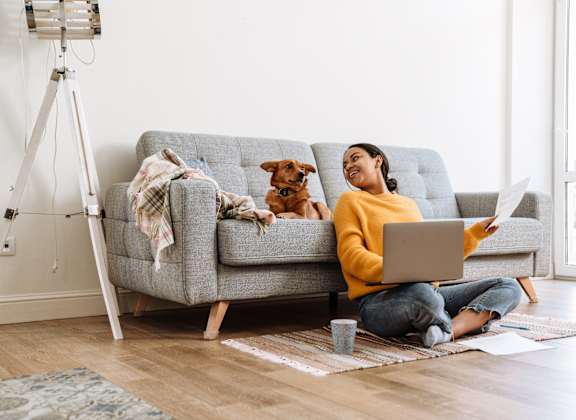 A woman sitting on the floor in front of a couch with a laptop, a cup, and some papers.