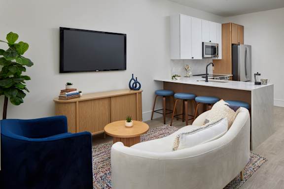 A living room with a white couch, a blue chair, a wooden cabinet, a TV, a microwave, a refrigerator, and a small table.