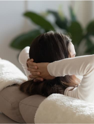A woman is sitting on a couch with her back to the camera, wearing a white long-sleeve shirt.