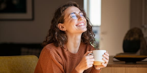 A woman with glasses is smiling while holding a cup of coffee.