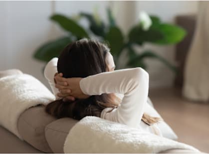 A woman is sitting on a couch with her back to the camera, wearing a white long-sleeve shirt.