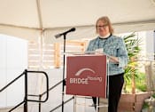 Thumbnail 19 of 24 - A woman stands behind a podium with a sign that says Bridge Housing.