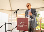 Thumbnail 18 of 24 - A man in a grey suit stands at a podium with a sign that says "BRIDGE HOUSING".