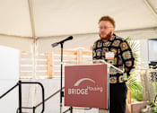 Thumbnail 20 of 24 - A man is giving a speech at a podium with a sign that says BRIDGE Housing.
