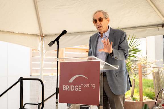 A man in a grey suit stands at a podium with a sign that says "BRIDGE HOUSING".