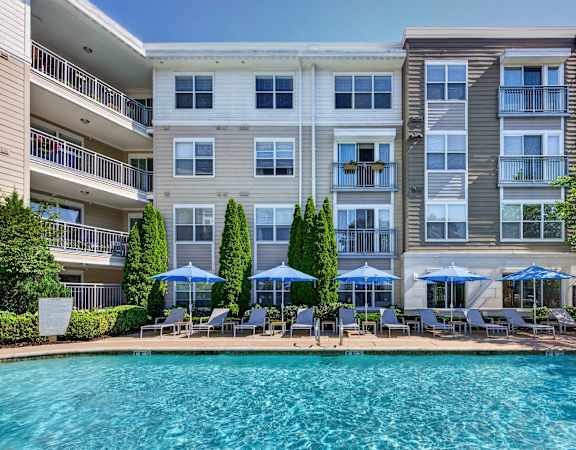 A large swimming pool is surrounded by lounge chairs and umbrellas in front of a multi-story apartment building.