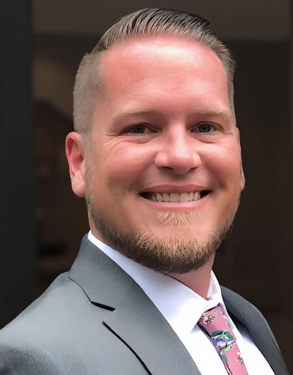 A man with a beard and short hair wearing a grey suit and a pink tie.