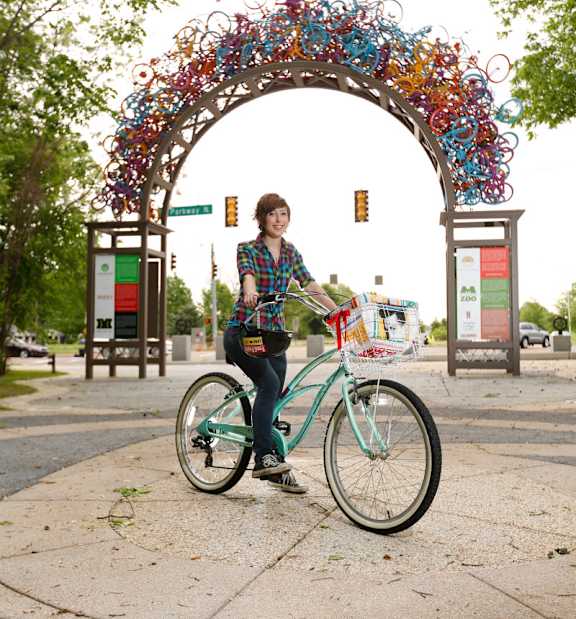 A woman is standing with a bicycle in front of a colorful arch.