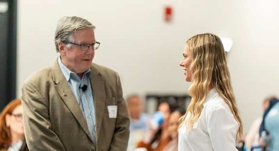A man in a brown jacket and a woman in a white shirt are having a conversation in a room with people sitting in the background.