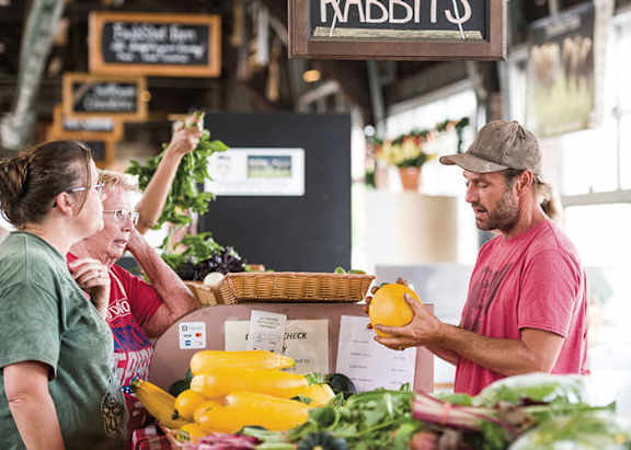 A man in a pink shirt is holding a lemon and talking to a woman in a green shirt at a market.
