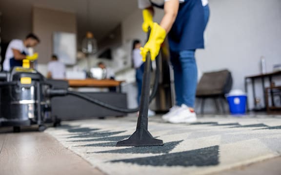 A person in a blue shirt and yellow gloves is vacuuming a rug in a room with other people in the background.