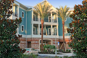An exterior view of a 3-story apartment building at The Flats at Tioga Town Center with patios or balconies and well-maintained palm trees near resident parking.