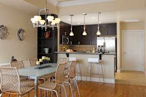 A modern kitchen with stainless steel appliances, hardwood-style flooring, pendant lights, a breakfast bar, and a built-in corner shelving unit near a dining area.
