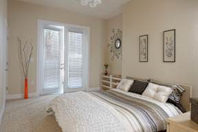 A staged bedroom with plush carpet flooring, ample natural light, and double doors to a private outdoor balcony.