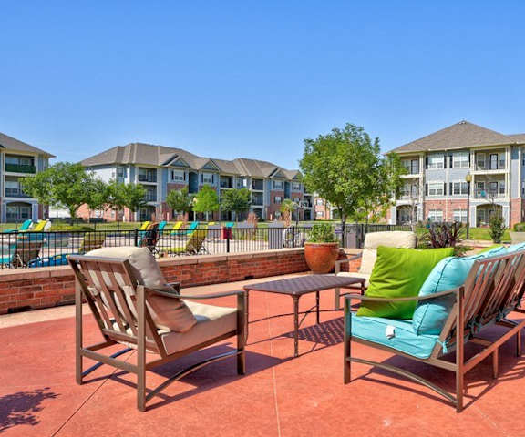The outdoor loounge area with colorful chairs and couches, as well as tables, with plants, the pool, and the buildings in the background at Liberty Pointe Apartment Homes. 