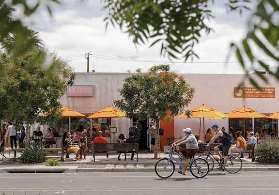 A group of people are sitting and standing outside of a restaurant with orange umbrellas.