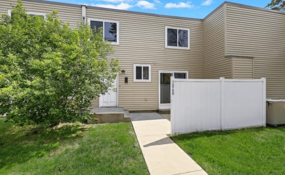 the back of a brown house with a sidewalk and a white fence