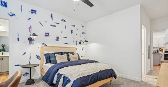 A bedroom with a bed, a ceiling fan, and a wall with blue paint splatters.