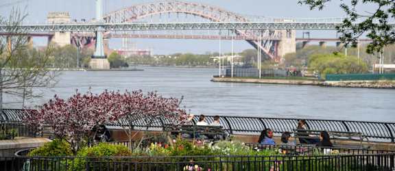 A bridge spans across a river with people sitting on a bench in the foreground.