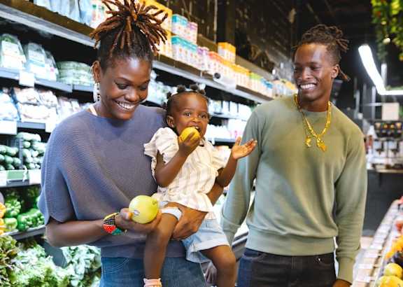A woman with a child in her arms is holding a lemon while standing next to a man in a grocery store.