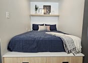 Thumbnail 7 of 7 - A bed with a blue comforter and pillows is in a room with a shelf above it.