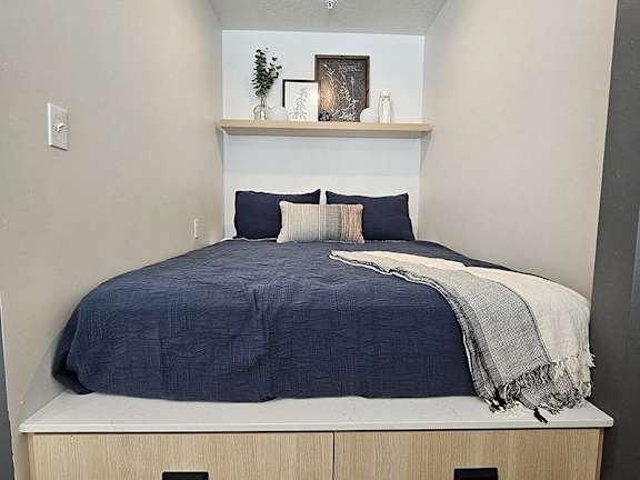 A bed with a blue comforter and pillows is in a room with a shelf above it.