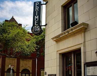 A white car is parked in front of a building with a sign that says "Prime 115.".