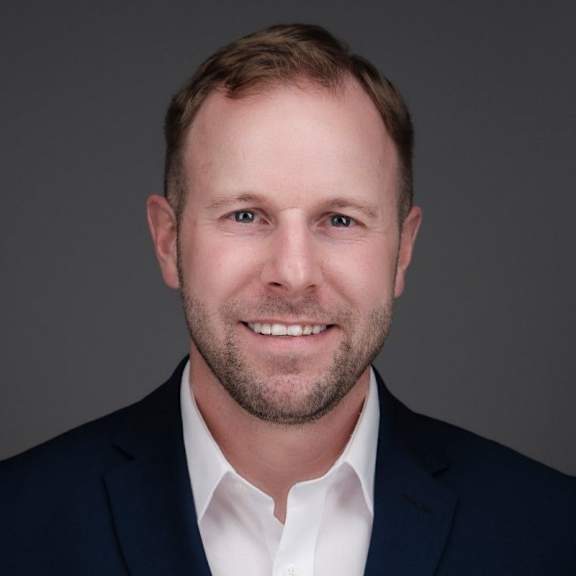 A man with a beard and short hair wearing a dark suit and white shirt.