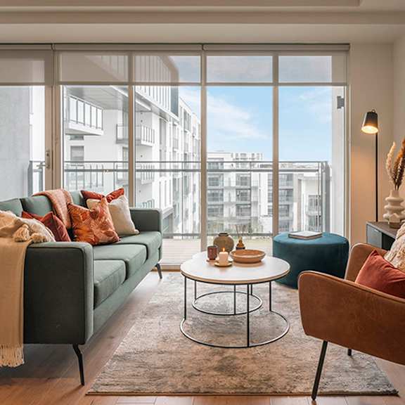 A living room with a green couch, a brown chair, a white table, and a large window with a view of buildings outside at Cherrywood Apartments, D18 VY3V, DUBLIN 18