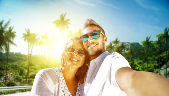 a man and a woman wearing sunglasses smile at the camera with palm trees in the background