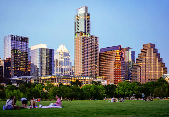 A group of people are sitting on a grassy hill with a city skyline in the background.