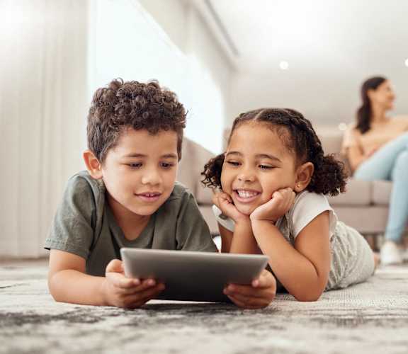 Two children are lying on the floor playing with a tablet.