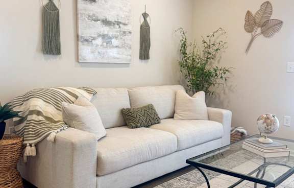 A living room with a white couch, a glass coffee table, and two wall hangings.