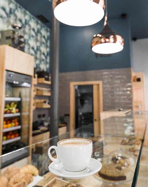 A cup of coffee is on a saucer in front of a glass display case filled with baked goods.
