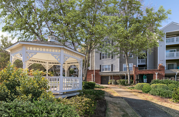 A white gazebo is situated in front of a building with a tree in the background.