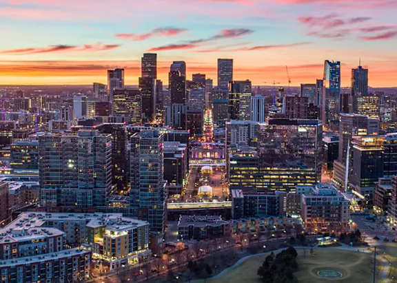 A cityscape at dusk with buildings illuminated and a park in the foreground.