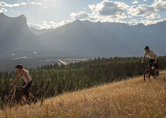 Two people riding bikes on a grassy hill with mountains in the background.