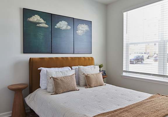A bedroom with a bed, pillows, and a painting of clouds on the wall.