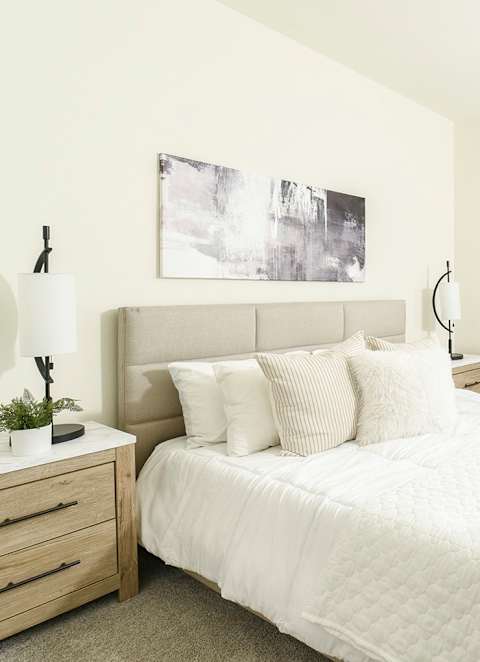 A white bedroom with a large bed, a nightstand, a table, and a window with blinds.