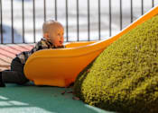 Thumbnail 2 of 7 - A baby is sitting in a yellow bathtub next to a green bush.