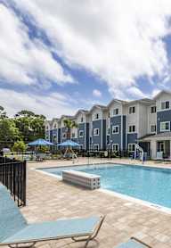A swimming pool is surrounded by a patio and chairs in front of apartment buildings.