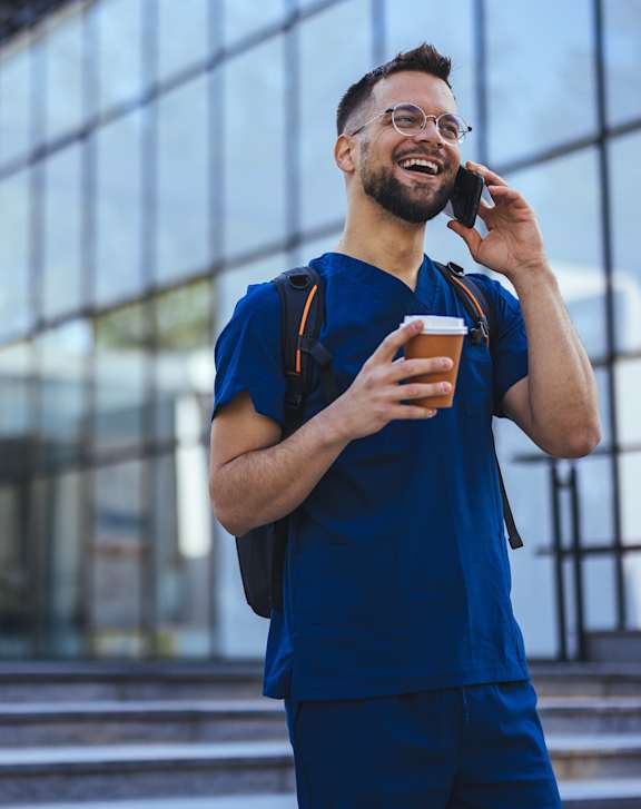 A man in blue scrubs is talking on a phone while holding a cup.