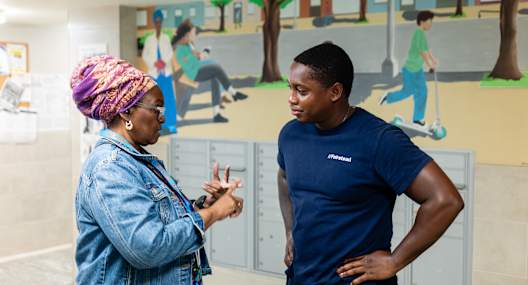 A woman in a denim jacket and a man in a blue shirt are standing in front of a wall with a mural of children playing.