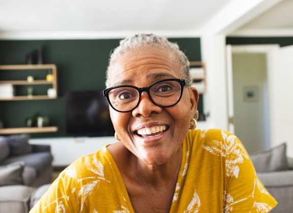A woman with glasses and a yellow top is smiling in a living room.