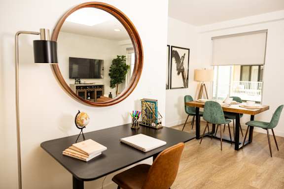 A room with a round mirror, a desk with a chair, a lamp, a plant, a globe, a book, and a potted plant.