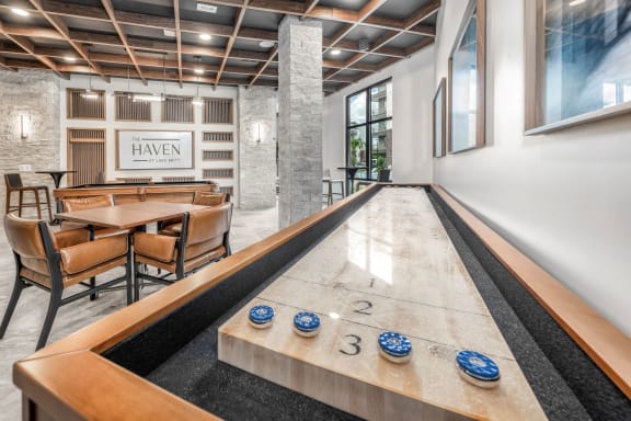 A shiny wooden table with blue and white coasters is in the foreground of a room with a bowling alley. at The Haven at Lake Britt Apartments, Winter Garden, FL 34787
