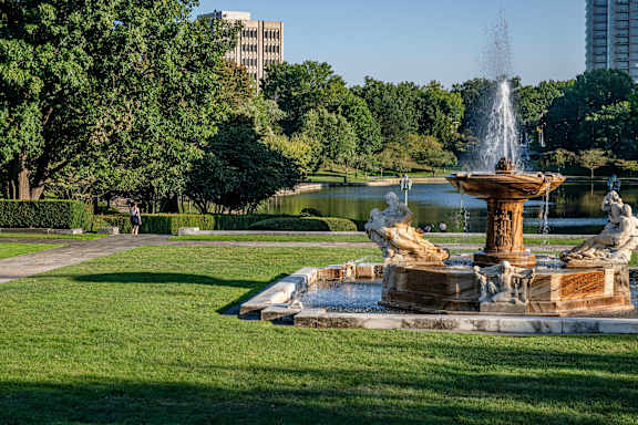 A fountain in the middle of a park with trees and buildings in the background.