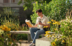 A man is sitting on a bench reading a book and holding a phone.