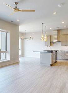 An airy open-concept living and kitchen area with light wood-style flooring, modern ceiling fan, floor-to-ceiling windows, pendant lighting over the island, and stainless steel appliances.